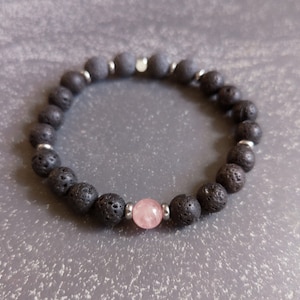 May include: A beaded bracelet featuring black lava rock beads, interspersed with small silver-colored spacers. A single, round, pink rose quartz bead is centered in the design. The bracelet is strung on an elastic cord.