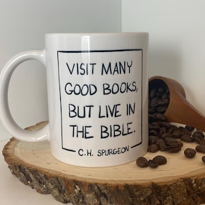 以下が含まれることがあります： 黒い文字で「VISIT MANY GOOD BOOKS, BUT LIVE IN THE BIBLE. C.H. SPURGEON」と書かれた白いセラミックマグ。マグカップは、コーヒー豆と木のスプーンが付いた木の輪切りに置かれています。