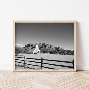 May include: Black and white framed photograph of a church with a steeple, set against a backdrop of mountains. A wooden fence is in the foreground. The image is displayed on a light-colored wooden floor.