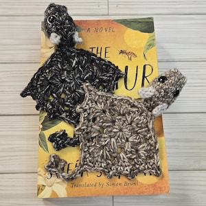 May include: Two handmade crochet bookmarks shaped like animals, one black and white, the other brown and white, rest on a book titled "The Fur". The book has a yellow cover with a floral design and the text "A Novel".