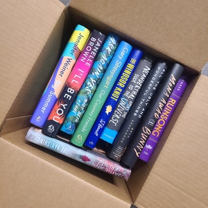 May include: A cardboard box filled with a stack of paperback books. The books are various colors and have titles such as "The Windsor Knot", "Ask Again, Yes", and "I'll Be You".