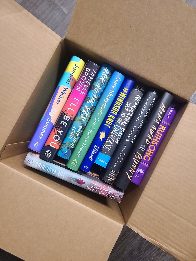 May include: A cardboard box filled with a stack of paperback books. The books are various colors and have titles such as "The Windsor Knot", "Ask Again, Yes", and "I'll Be You".