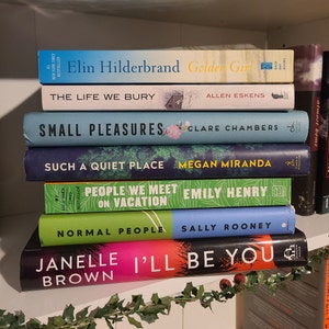 May include: A stack of paperback books on a white shelf with green garland. The books are all different colors and have various titles, including "Golden Girl", "Small Pleasures", "Such a Quiet Place", "People We Meet on Vacation", "Normal People", and "I'll Be You".