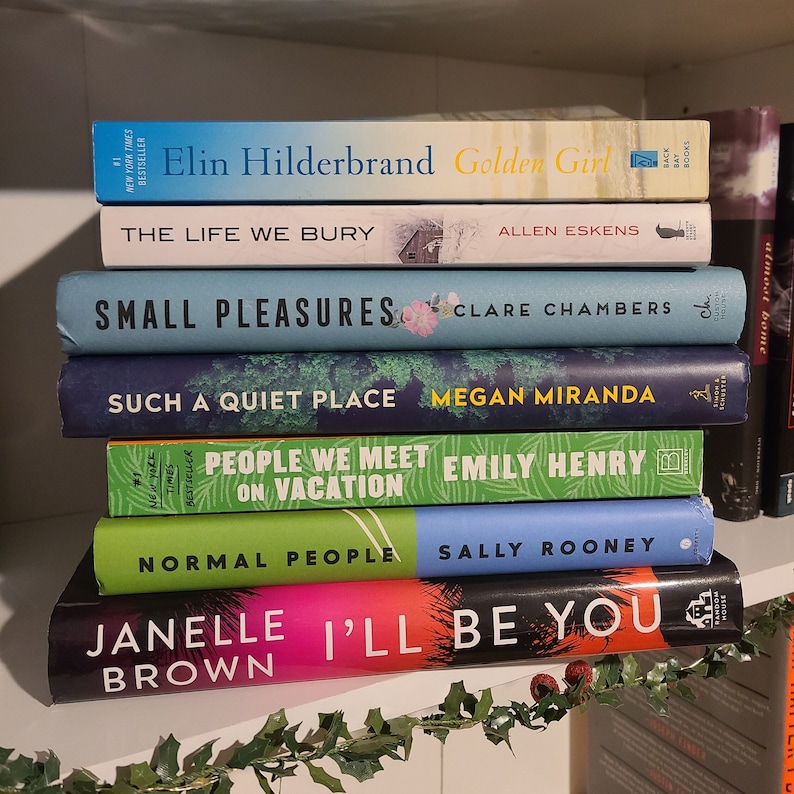 May include: A stack of paperback books on a white shelf with green garland. The books are all different colors and have various titles, including "Golden Girl", "Small Pleasures", "Such a Quiet Place", "People We Meet on Vacation", "Normal People", and "I'll Be You".