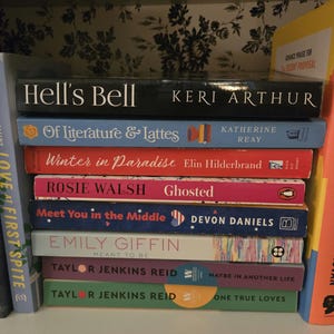 May include: A bookshelf with a variety of paperback books stacked on top of each other. The books are all different colors and have different titles. Some of the titles include "Hell's Bell", "Of Literature & Lattes", "Winter in Paradise", "Ghosted", "Meet You in the Middle", "Meant to Be", "Maybe in Another Life", and "One True Loves".