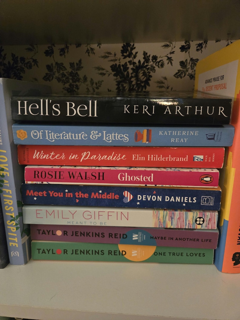 May include: A bookshelf with a variety of paperback books stacked on top of each other. The books are all different colors and have different titles. Some of the titles include "Hell's Bell", "Of Literature & Lattes", "Winter in Paradise", "Ghosted", "Meet You in the Middle", "Meant to Be", "Maybe in Another Life", and "One True Loves".
