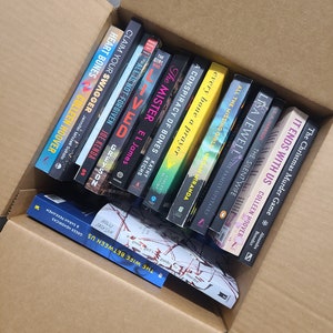 May include: A cardboard box filled with a variety of paperback books. The books are stacked vertically and include titles such as "The Wife Between Us", "The Christmas Murder Game", "All the Missing Girls", and "The Silent Wife".