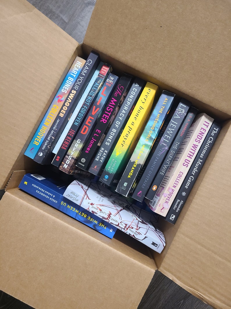 May include: A cardboard box filled with a variety of paperback books. The books are stacked vertically and include titles such as "The Wife Between Us", "The Christmas Murder Game", "All the Missing Girls", and "The Silent Wife".