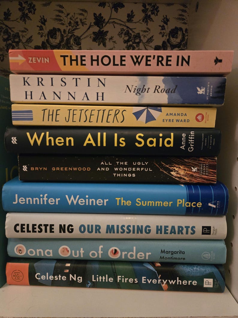 May include: A stack of ten paperback books with blue, green, yellow, and white covers. The titles of the books are visible: "The Hole We're In", "Night Road", "The Jetsetters", "When All Is Said", "All the Ugly and Wonderful Things", "The Summer Place", "Our Missing Hearts", "Oona Out of Order", and "Little Fires Everywhere".