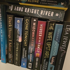 May include: A bookshelf with a collection of paperback books, including "You Are Not Alone", "Long Bright River", "The Next Mrs. Parrish", "Dark Things I Adore", "Imaginary Friend", "Gone Tonight", "Widowmaker", "Murder in the Family", "The Wife Upstairs", and "The Family Upstairs".