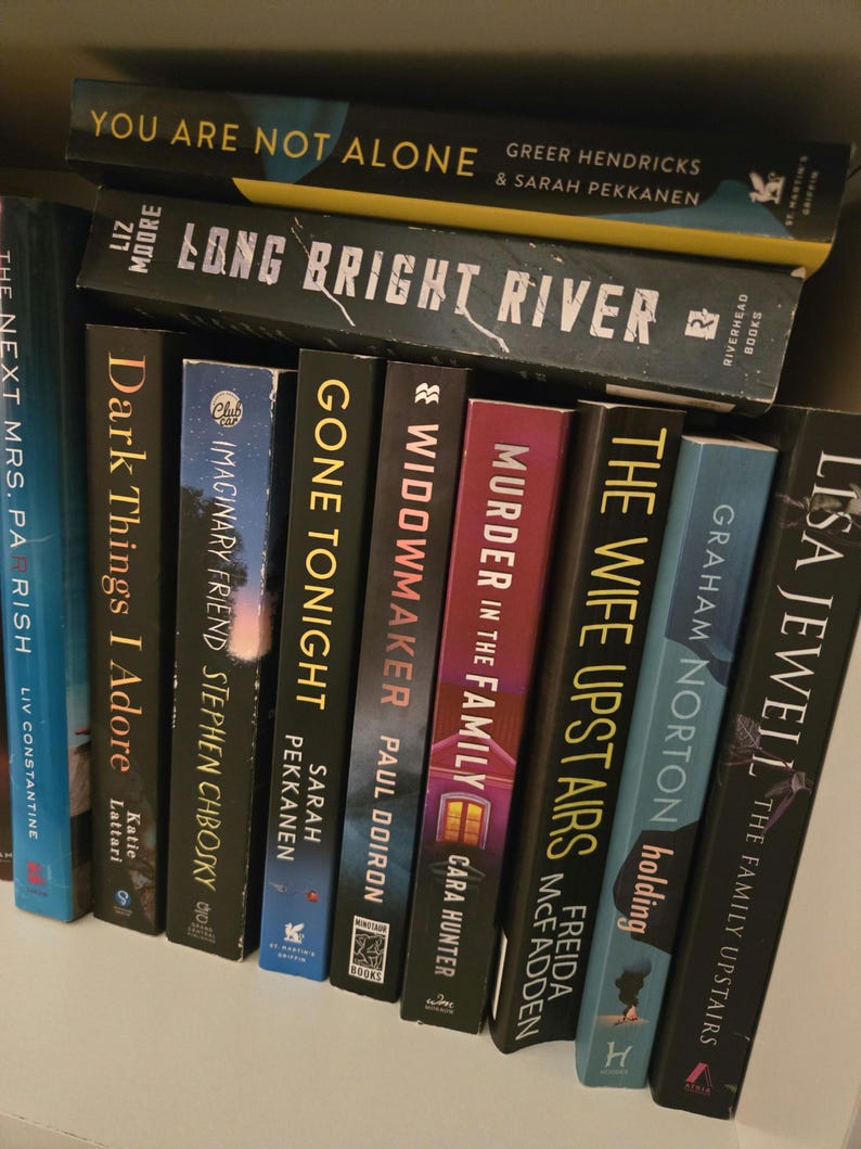 May include: A bookshelf with a collection of paperback books, including "You Are Not Alone", "Long Bright River", "The Next Mrs. Parrish", "Dark Things I Adore", "Imaginary Friend", "Gone Tonight", "Widowmaker", "Murder in the Family", "The Wife Upstairs", and "The Family Upstairs".
