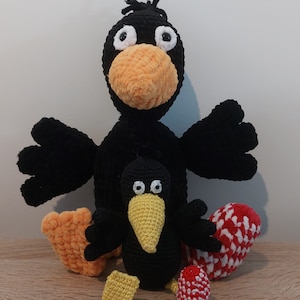 Könnte beinhalten: Zwei schwarze gehäkelte Krähen, eine große und eine kleine, mit orangefarbenen und gelben Schnäbeln und weißen Augen. Die größere Krähe hat rote und weiße gehäkelte Eier daneben.