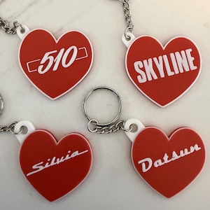 May include: Red heart-shaped keyrings with white text. The keyrings display the words "510", "Skyline", "Silvia", and "Datsun". Each keyring has a silver chain and a ring.