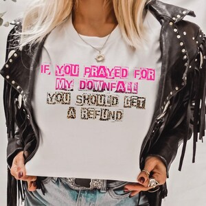 May include: White t-shirt with the text "IF YOU PRAYED FOR MY DOWNFALL YOU SHOULD GET A REFUND" in pink and leopard print. The shirt is worn with a black leather jacket with fringe and silver studs.