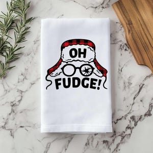 May include: White kitchen towel with a red and black plaid design of a character wearing a hat and glasses with the text "OH FUDGE!"