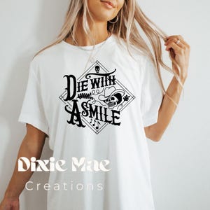 May include: White t-shirt with a black graphic design that says "Die with a Smile" and features a skull wearing a cowboy hat. The text "Dixie Mae Creations" is printed below the graphic.