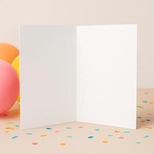 May include: An open, blank greeting card with a white interior, standing upright on a light beige surface. Colorful confetti is scattered around the card. Balloons in shades of pink, yellow, and orange are visible in the background.