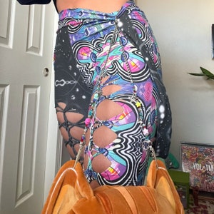 May include: A pair of leggings with a vibrant, psychedelic pattern in shades of purple, blue, and pink. The leggings feature cut-out details with a lace-up design and are adorned with a beaded chain. The overall theme is a unique and artistic fashion statement.