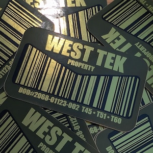 Puede incluir: Varias pegatinas rectangulares verde oliva con texto dorado y diseños de códigos de barras están dispersas sobre una superficie. Las pegatinas dicen "WEST TEK PROPERTY" e incluyen una serie de números y códigos. La estética general es industrial.