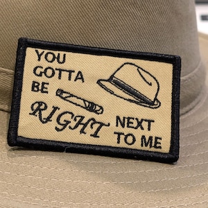 Könnte beinhalten: Beigefarbener Safarihut mit rechteckigem Aufnäher. Der Aufnäher zeigt eine Fedora und eine Zigarrenillustration mit dem Text "YOU GOTTA BE RIGHT NEXT TO ME" in schwarzer Schrift.