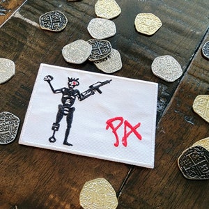 May include: White embroidered patch with a black skeleton holding a gun and the red text "PAX". The patch is surrounded by gold and silver pirate coins.