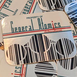 May include: A collection of metallic coasters with the text "PROPERTY OF General Atomics International." The coasters have a white background with a red and green border. Each coaster features a unique barcode design.