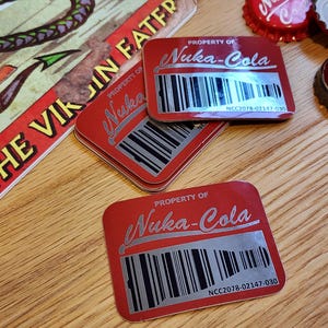 Może przedstawiać: Trzy czerwone prostokątne naklejki z tekstem "PROPERTY OF Nuka-Cola" i kodem kreskowym. Tekst "NCC2078-02147-030" jest nadrukowany pod kodem kreskowym na każdej naklejce.