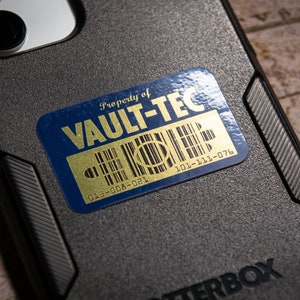 K&ouml;nnte beinhalten: Ein blauer und goldener Aufkleber mit dem Text "Property of VAULT-TEC" und Barcodes mit den Nummern "013-008-021" und "101-111-076".