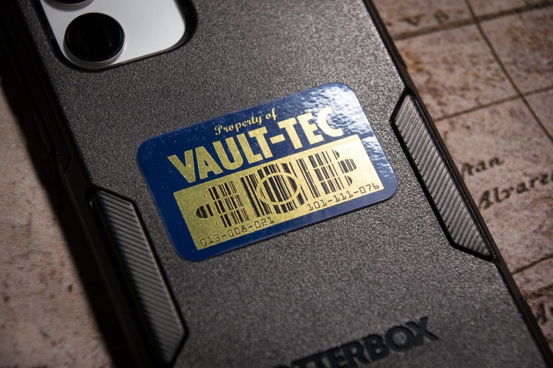 K&ouml;nnte beinhalten: Ein blauer und goldener Aufkleber mit dem Text "Property of VAULT-TEC" und Barcodes mit den Nummern "013-008-021" und "101-111-076".