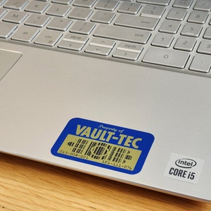 K&ouml;nnte beinhalten: Ein silberner Laptop mit einem blauen und goldenen Aufkleber, auf dem "Property of VAULT-TEC" steht, und einem schwarzen und silbernen Aufkleber, auf dem "(intel) CORE i5" steht.
