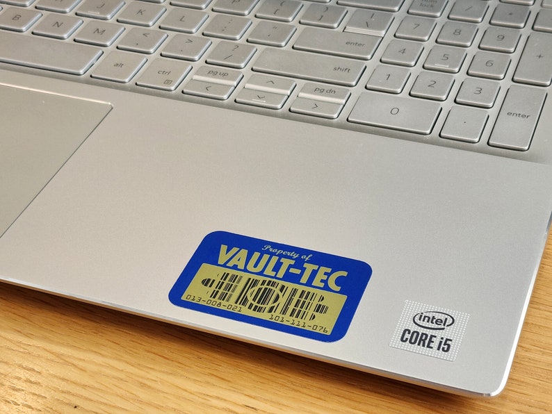 K&ouml;nnte beinhalten: Ein silberner Laptop mit einem blauen und goldenen Aufkleber, auf dem "Property of VAULT-TEC" steht, und einem schwarzen und silbernen Aufkleber, auf dem "(intel) CORE i5" steht.