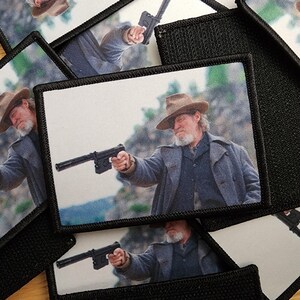 Könnte beinhalten: Rechteckige Aufnäher mit einem Mann mit Hut, der eine Waffe anvisiert. Der Mann trägt einen langen Mantel und einen Hut. Die Aufnäher haben einen schwarzen Rand und eine schwarze Stoffrückseite. Das Bild ist eine Nahaufnahme der Aufnäher.