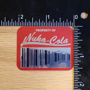 Może przedstawiać: Czerwona naklejka z tekstem "PROPERTY OF Nuka-Cola" w srebrnych literach. Pod tekstem znajduje się czarno-biały kod kreskowy z numerem "NCC2078 02147-030" wydrukowanym pod kodem kreskowym.