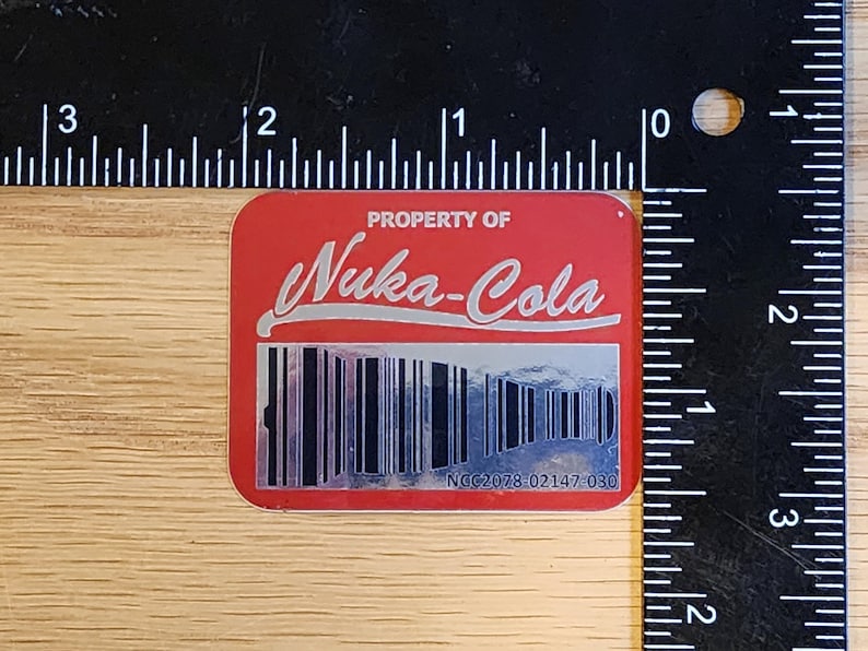 Może przedstawiać: Czerwona naklejka z tekstem "PROPERTY OF Nuka-Cola" w srebrnych literach. Pod tekstem znajduje się czarno-biały kod kreskowy z numerem "NCC2078 02147-030" wydrukowanym pod kodem kreskowym.
