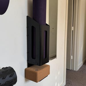 May include: A black metal wall-mounted yoga mat holder containing a rolled purple yoga mat. A brown cork yoga block is positioned below. The holder is fixed to a white wall.