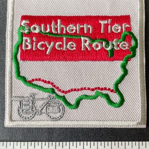 Op de afbeelding: Witte geborduurde patch met een groene omtrek van de Verenigde Staten en een rode lijn die de Southern Tier Bicycle Route voorstelt. De tekst "Southern Tier Bicycle Route" staat in wit op een rode achtergrond.