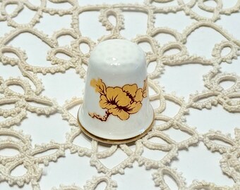 May include: A white porcelain thimble with a gold trim and a floral design featuring a yellow flower with brown leaves.