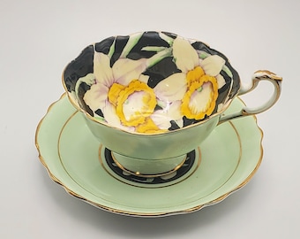 May include: A vintage teacup and saucer set with a black background and a floral design of white and yellow daffodils. The saucer is a pale green color.
