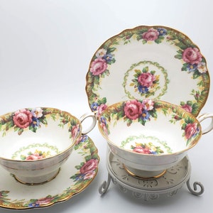 Puede incluir: Un juego de tres tazas de té y platillos vintage con un patrón floral. Las tazas y los platillos son blancos con un borde dorado y presentan un delicado diseño floral en tonos de rosa, azul y verde.