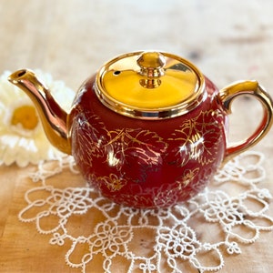 May include: A red teapot with gold leaf detailing and a gold lid. The teapot is sitting on a white crocheted doily.