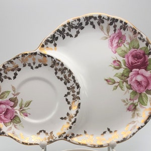 May include: White porcelain serving dish with a scalloped edge, featuring a floral rose design in pink and green. The rim is decorated with black and gold leaf patterns. A small, round indentation is on the left side.