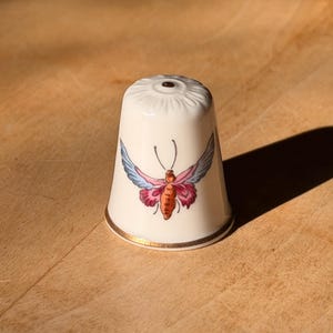 May include: A white porcelain thimble with a gold rim and a colorful butterfly design. The butterfly has pink, blue, and orange wings. The thimble is sitting on a wooden surface.