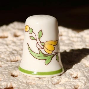 May include: A white ceramic thimble with a floral design. The thimble features a yellow flower with green leaves and stems. A green stripe encircles the base. The thimble is likely used for sewing or crafting.