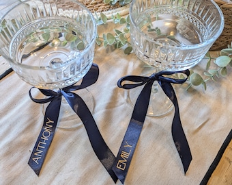 May include: Two clear glass champagne flutes with black ribbon bows tied around the stems. The ribbons are labeled "Anthony" and "Emily" in gold lettering.