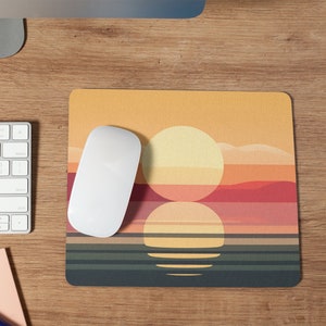 May include: A sunset-themed mousepad with a white mouse on top. The mousepad features a stylized sunset with a yellow, orange, and red sky, and a dark blue ocean with three horizontal lines representing waves.