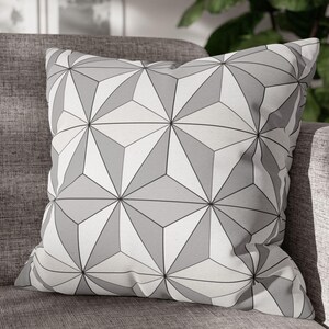 May include: Square throw pillow with a geometric pattern of gray and white triangles. The pillow is resting on a gray sofa, offering a modern decorative accent for home decor.