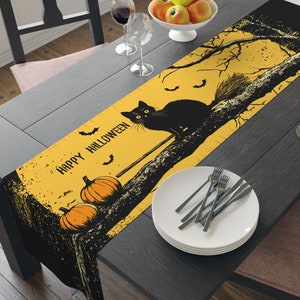 May include: A black and yellow Halloween table runner with a black cat sitting on a branch with a broom. The text "Happy Halloween" is printed on the runner.
