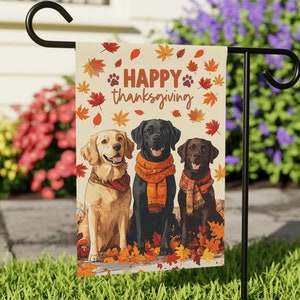 Pode incluir: Uma bandeira de jardim com três cães usando cachecóis e cercados por folhas de outono. A bandeira diz "Feliz Dia de Ação de Graças".