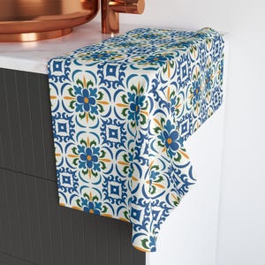 May include: A decorative hand towel with a blue, green, and yellow floral pattern on a white background. The towel is draped over a countertop near a copper sink and faucet. The pattern features stylized flowers and geometric shapes.