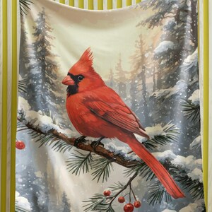 Könnte beinhalten: Eine Decke mit einem leuchtend roten Kardinal, der auf einem schneebedeckten Ast mit roten Beeren sitzt. Der Hintergrund zeigt eine verschneite Waldszene mit Nadelbäumen. Die Decke wird vor einer gelb-weiß gestreiften Wand gehalten.